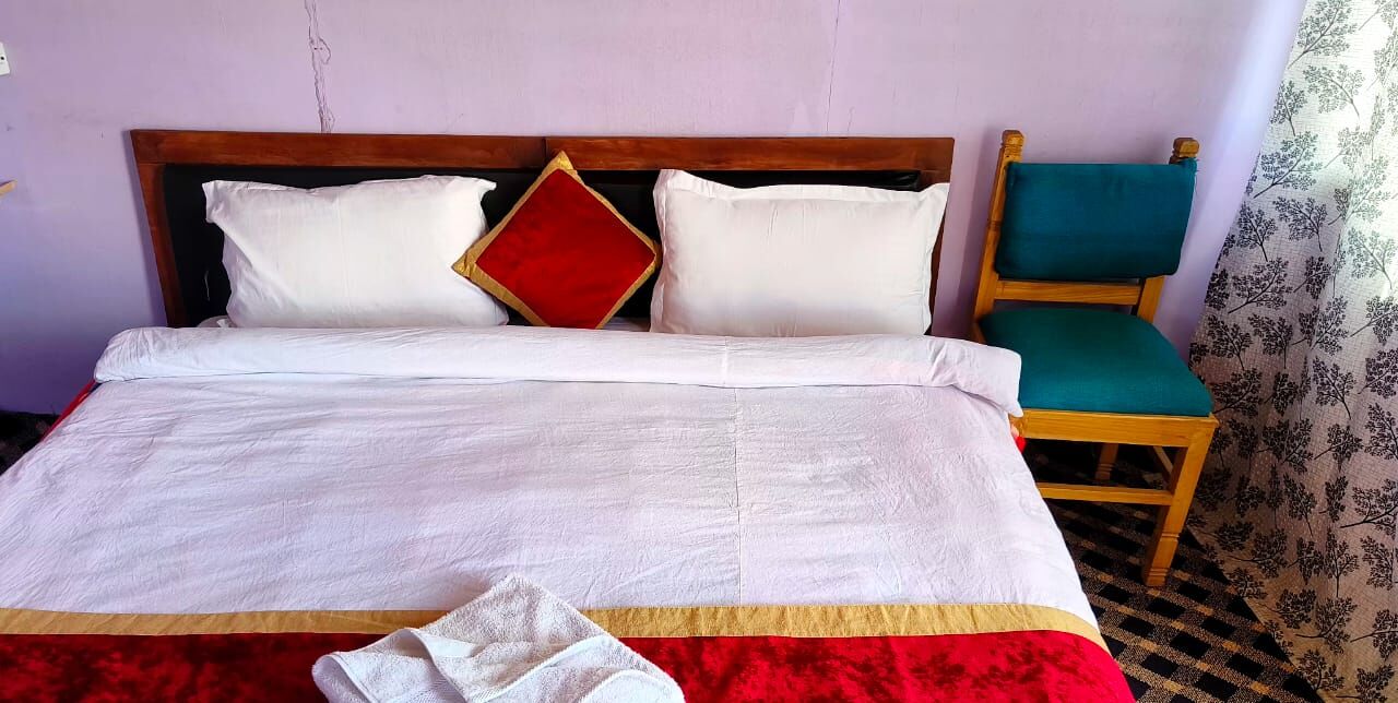 A neatly made bed with white bedding and a red accent pillow, a wooden headboard, and a wooden chair with a green cushion.