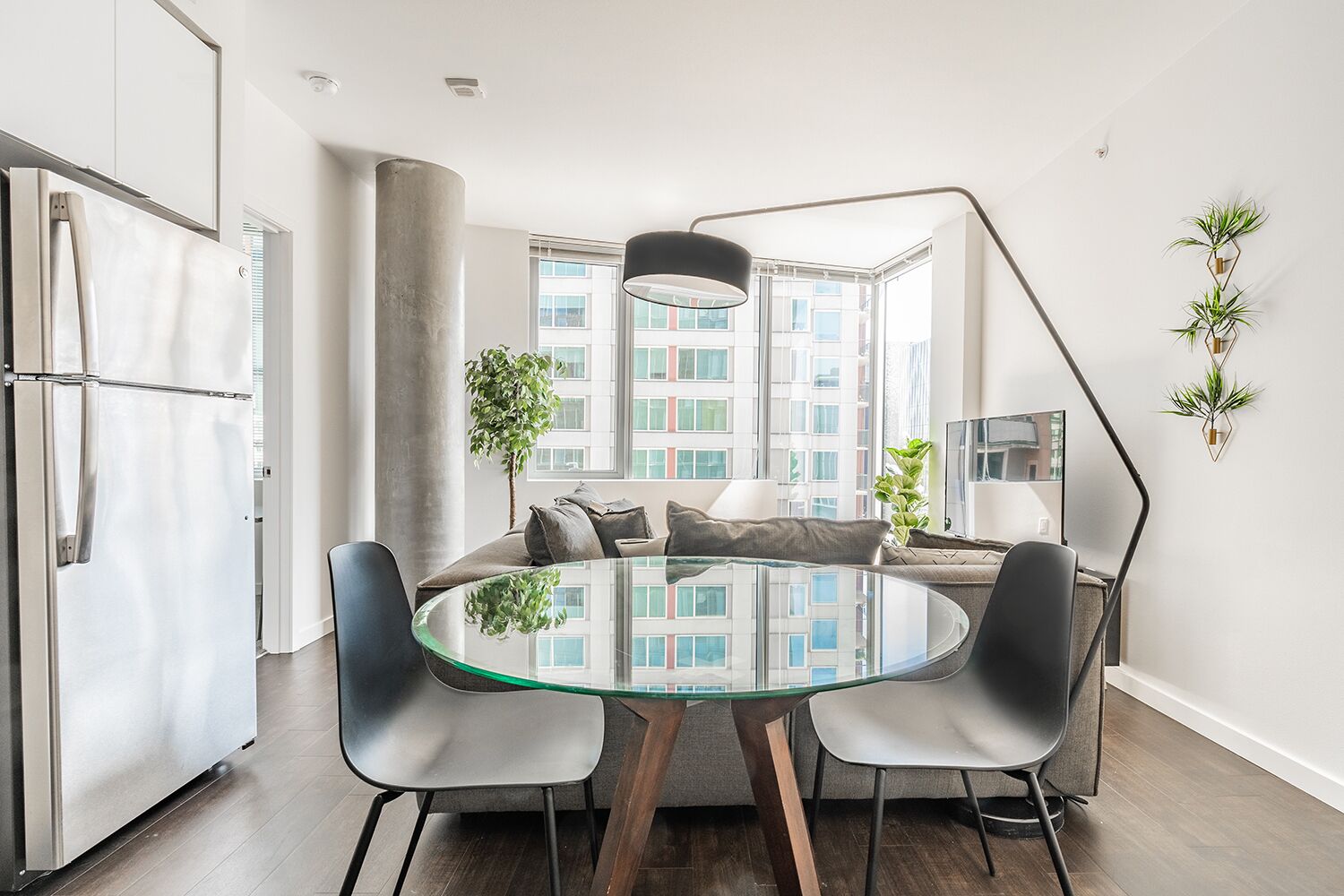Une cuisine moderne avec une table à manger en verre, deux chaises, un réfrigérateur et une grande fenêtre donnant sur des immeubles.