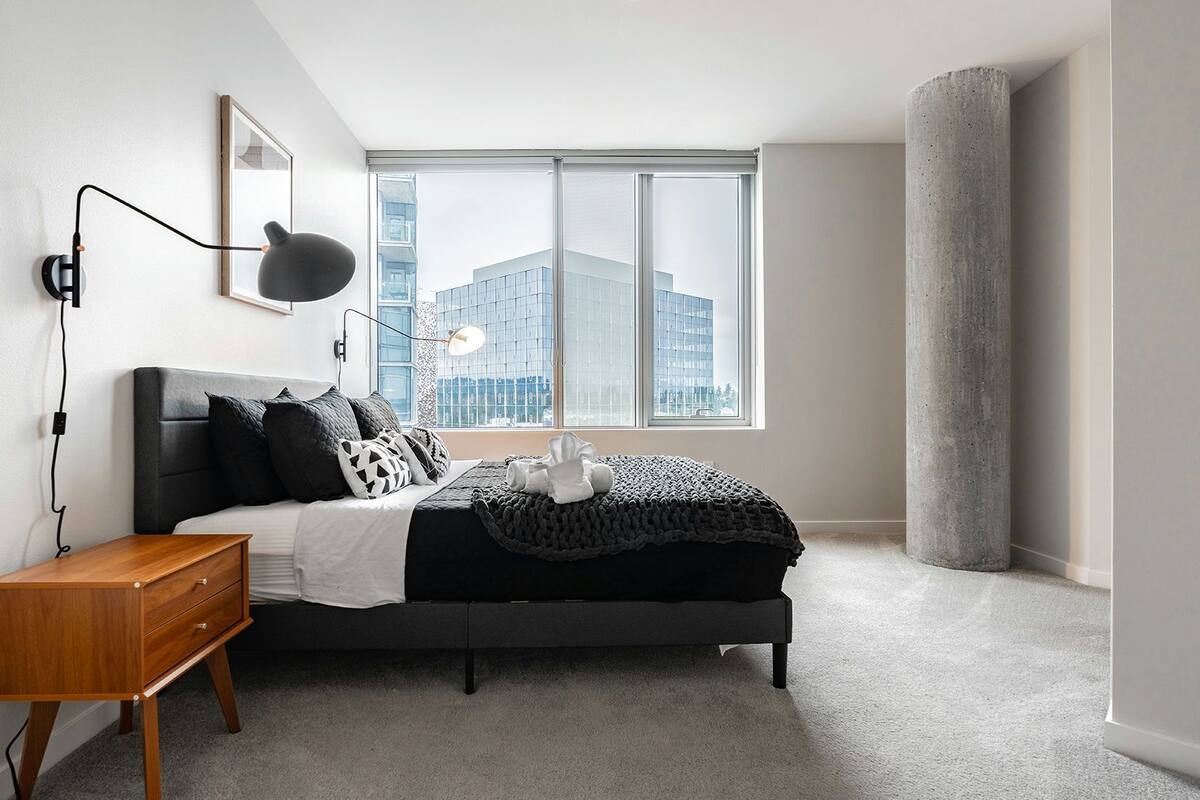 A modern bedroom with a bed, bedside table, lamp, and a large window with a city view.