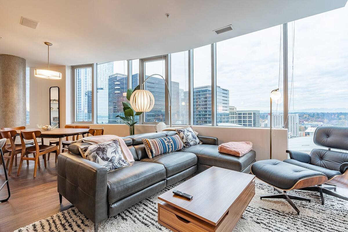 A modern living room with a sectional sofa, a coffee table, and a dining area with chairs and a table. Large windows offer a view of the cityscape.