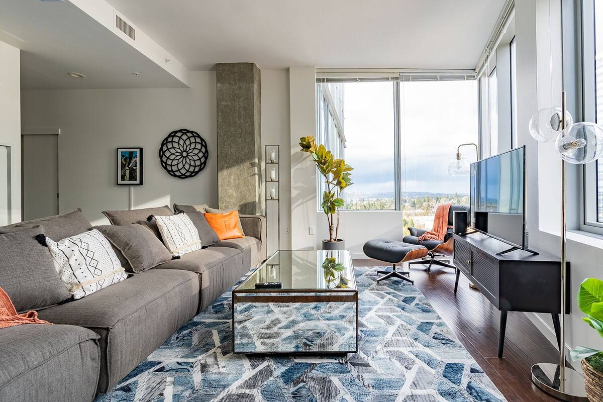 A modern living room with a grey sofa, a glass coffee table, and a large window offering a city view.