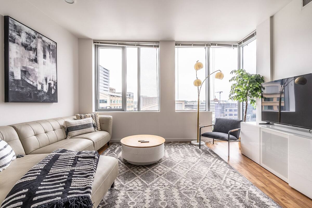 A modern living room with a sofa, a round coffee table, a TV, and large windows offering city views.