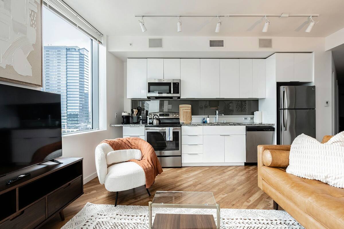 A modern kitchen with white cabinets, stainless steel appliances, and a large window overlooking a cityscape.