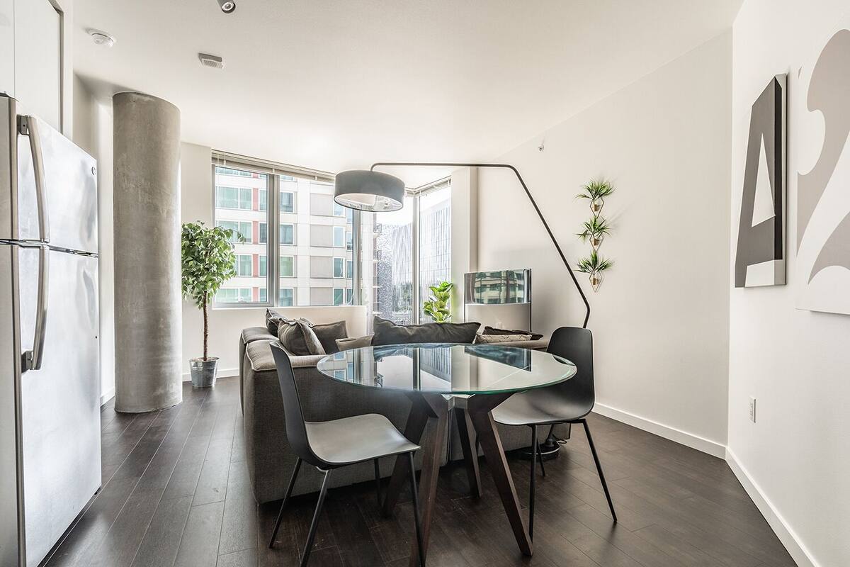 A modern kitchen with a round glass table, dark wooden flooring, and a large window offering a city view.