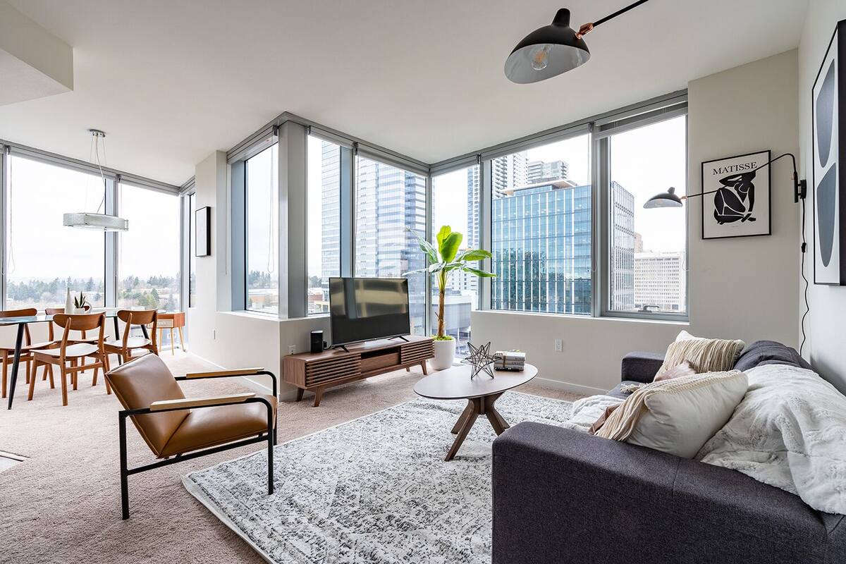 A modern living room with a large TV, a sofa, a dining area with chairs, and large windows offering a city view.