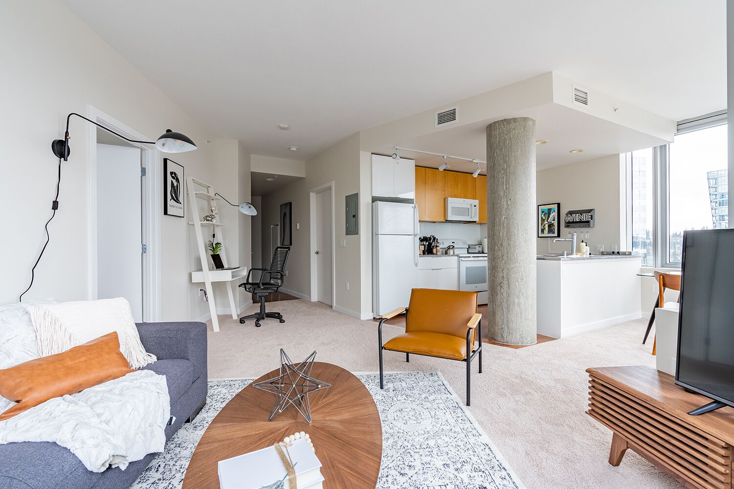 Una sala moderna con un sofá, una mesa de centro y un televisor. Hay una zona de cocina con un microondas y armarios.