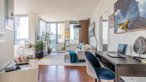 A modern living room with a sofa, a coffee table, and a desk with a laptop. There are large windows offering a view of the city. - Sophari Towers: Bellevue Apartments  (Bellevue)