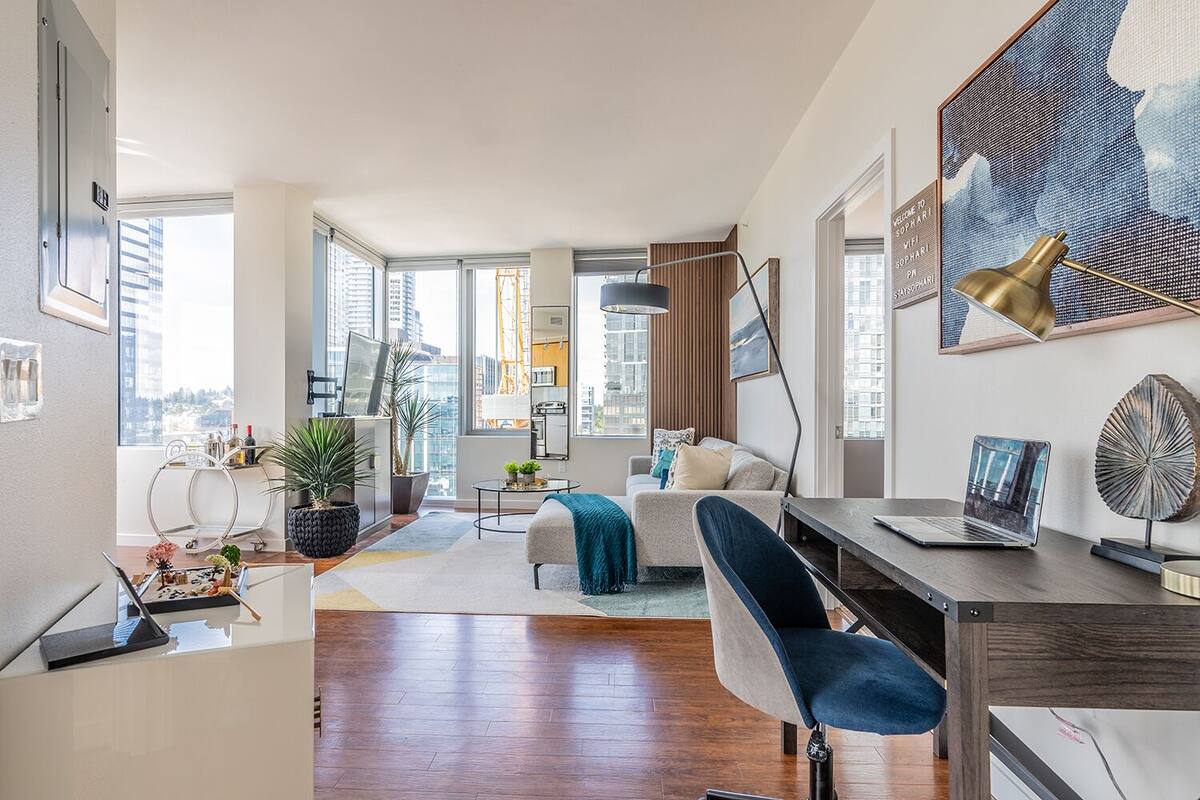 A modern living room with a sofa, coffee table, and a desk with a laptop. There is a large window with a view of buildings.