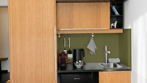 Une cuisine compacte avec des meubles en bois, un réfrigérateur noir, un évier et un petit plan de travail sur lequel se trouvent une bouilloire et un sac.