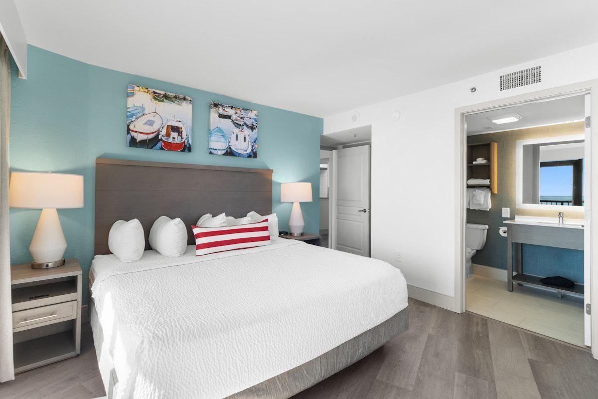 Quarto de hotel com uma cama grande, duas mesas de cabeceira com luminárias, cabeceira e uma obra de arte na parede.