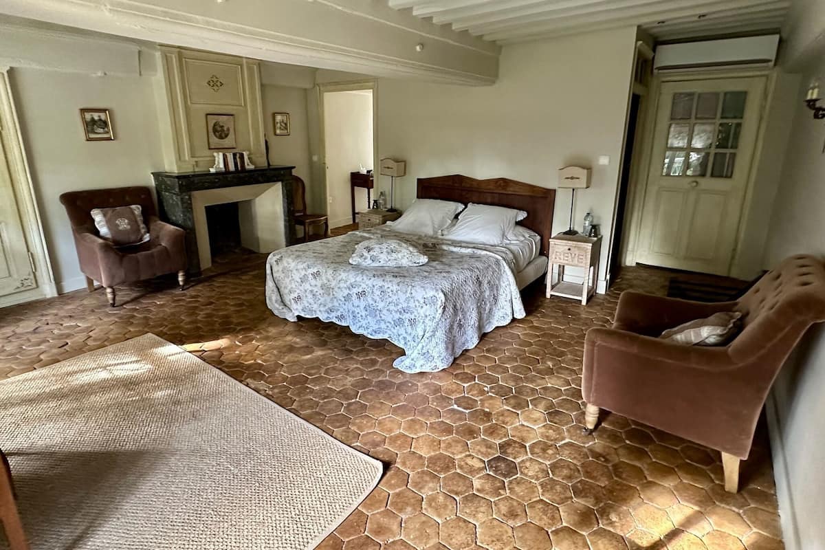 A bedroom with a large bed, a chair, a small table, and a fireplace.