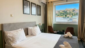 A hotel room with a large bed, a desk, two chairs, a window with a view of a historic building, and framed pictures on the wall.