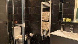 A modern bathroom with a glass shower enclosure, a white toilet, a towel rack with towels, a sink with a mirror, and dark tiled walls.