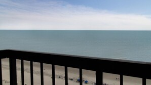 A beach view from a balcony with a railing, sandy beach, and a clear sky.