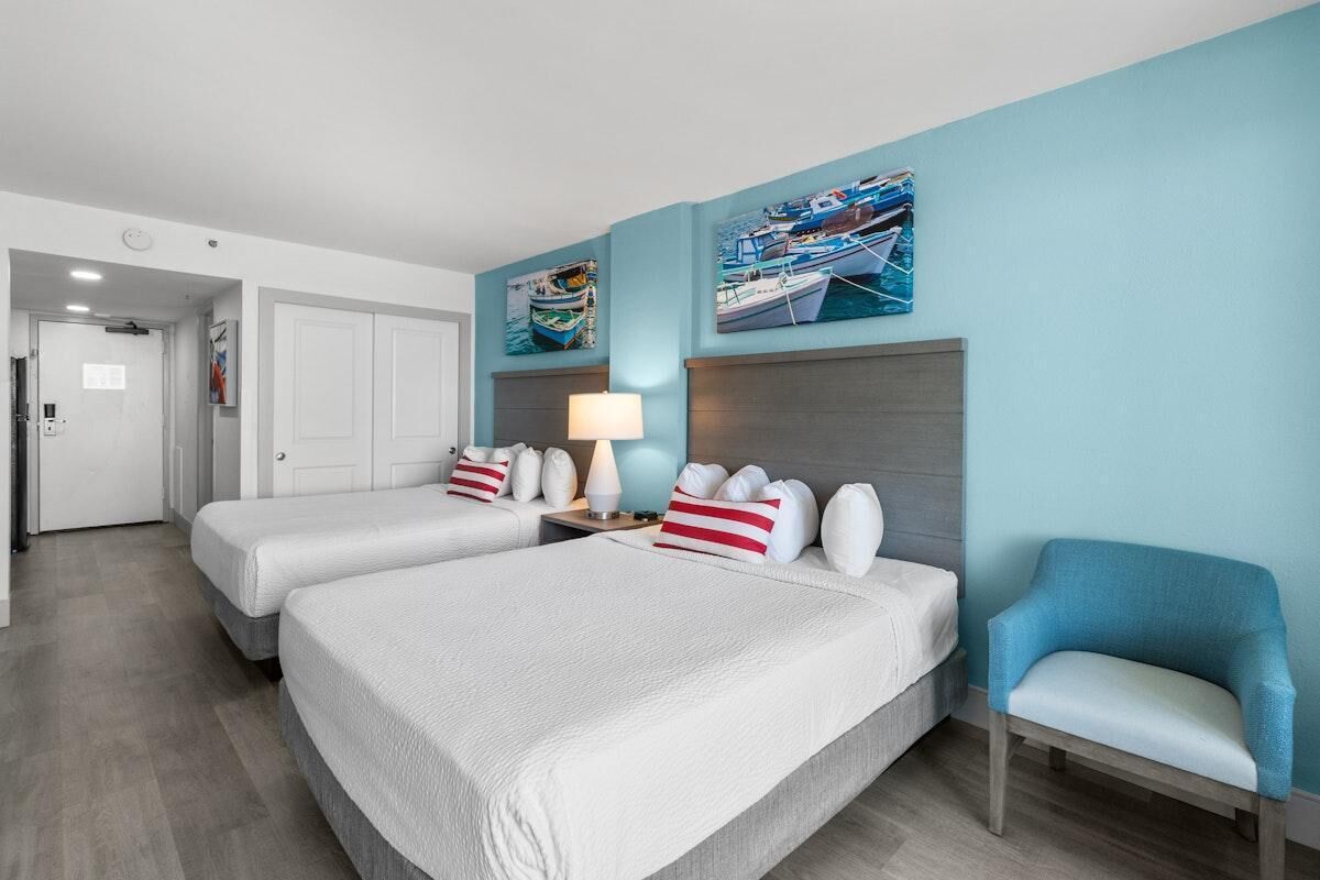 Een hotelkamer met twee bedden, een nachtkastje met lamp en een stoel. Twee schilderijen aan de muur beelden boten af.