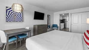 Habitación de hotel con cama, escritorio con sillas, televisor, espejo y zona de cocina visible a través de una puerta abierta.
