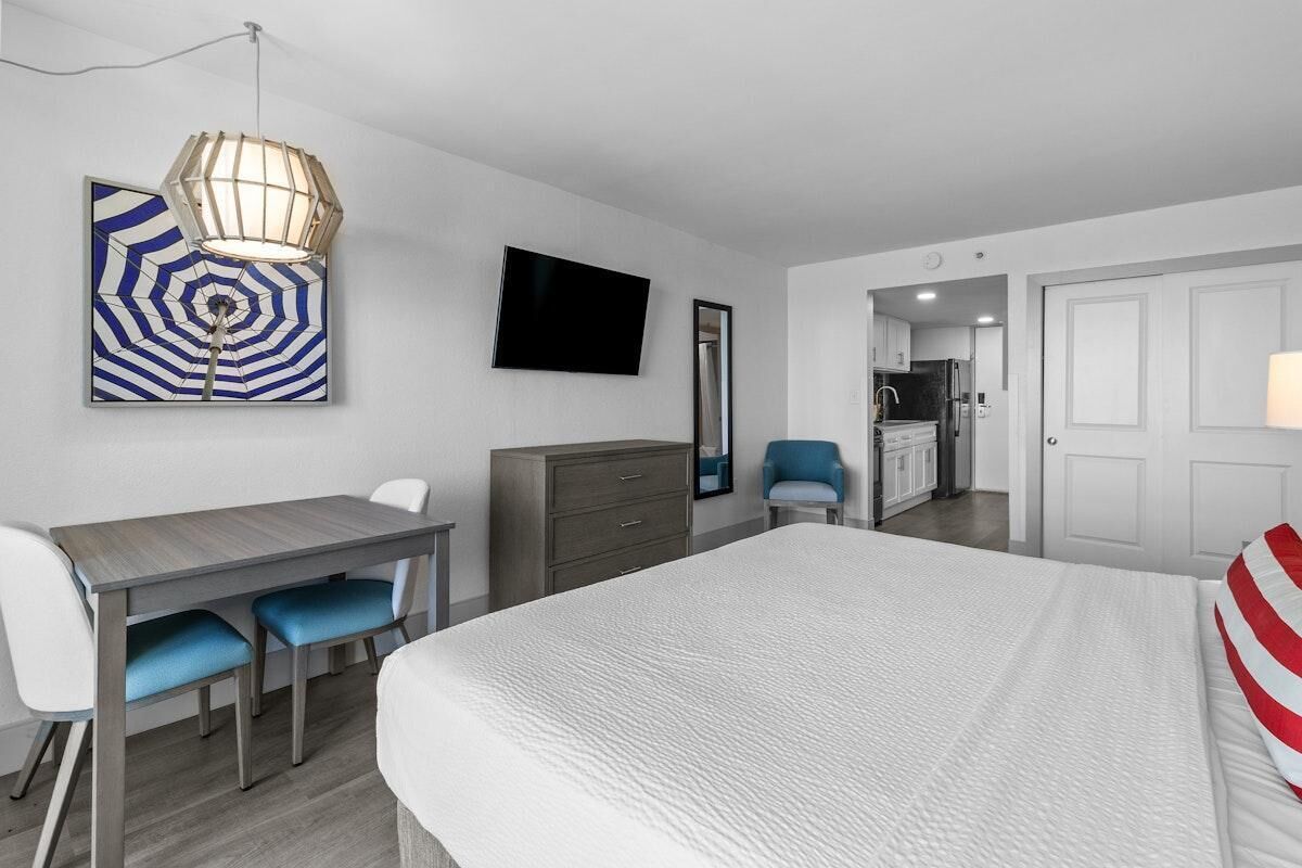 Ein Hotelzimmer mit Bett, Schreibtisch mit Stühlen, Fernseher, Spiegel und einer sichtbaren Küchenzeile durch eine offene Tür.