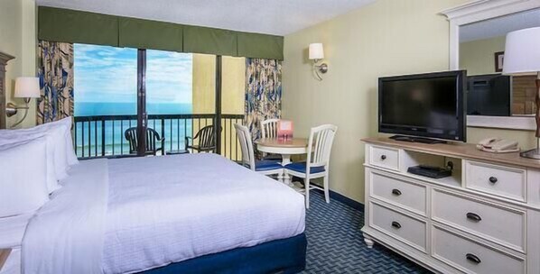 A hotel room with a bed, a TV, a dresser, a desk with a chair, and a view of the ocean. - Sunny Ocean View King Bed Accessible 5th Floor (Myrtle Beach)