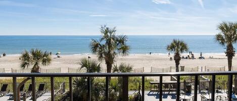A beach view from a balcony with a pool and lounge chairs.