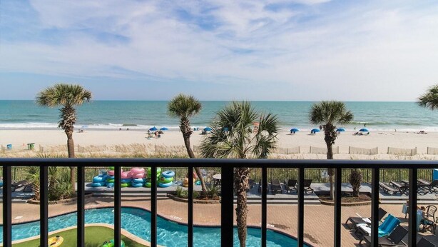 A pool area with lounge chairs and umbrellas, a beach with palm trees, and a view of the ocean. - Seaside Excellence Deluxe Room 4th Floor (Myrtle Beach)