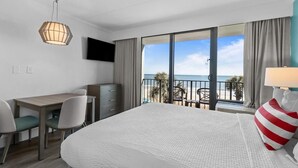 A hotel room with a bed, a desk, a chair, a TV, and a balcony with a view of the beach. - Southern Comfort Oceanfront Jr Suite 3rd Floor (Myrtle Beach)