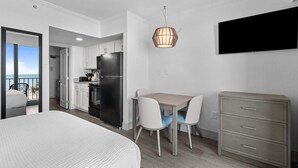 A bedroom with a bed, a dining area with a table and chairs, a kitchenette, a TV mounted on the wall, and a balcony with a view. - Extravagance Oceanfront Jr Suite 3rd Floor (Myrtle Beach)