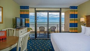 Et hotelværelse med seng, skrivebord med fjernsyn, stol, bord og udsigt til stranden gennem en skydedør.