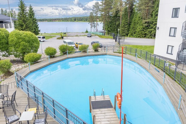 Une piscine avec une terrasse en bois, une bouée de sauvetage et une vue sur un lac et des bâtiments en arrière-plan.