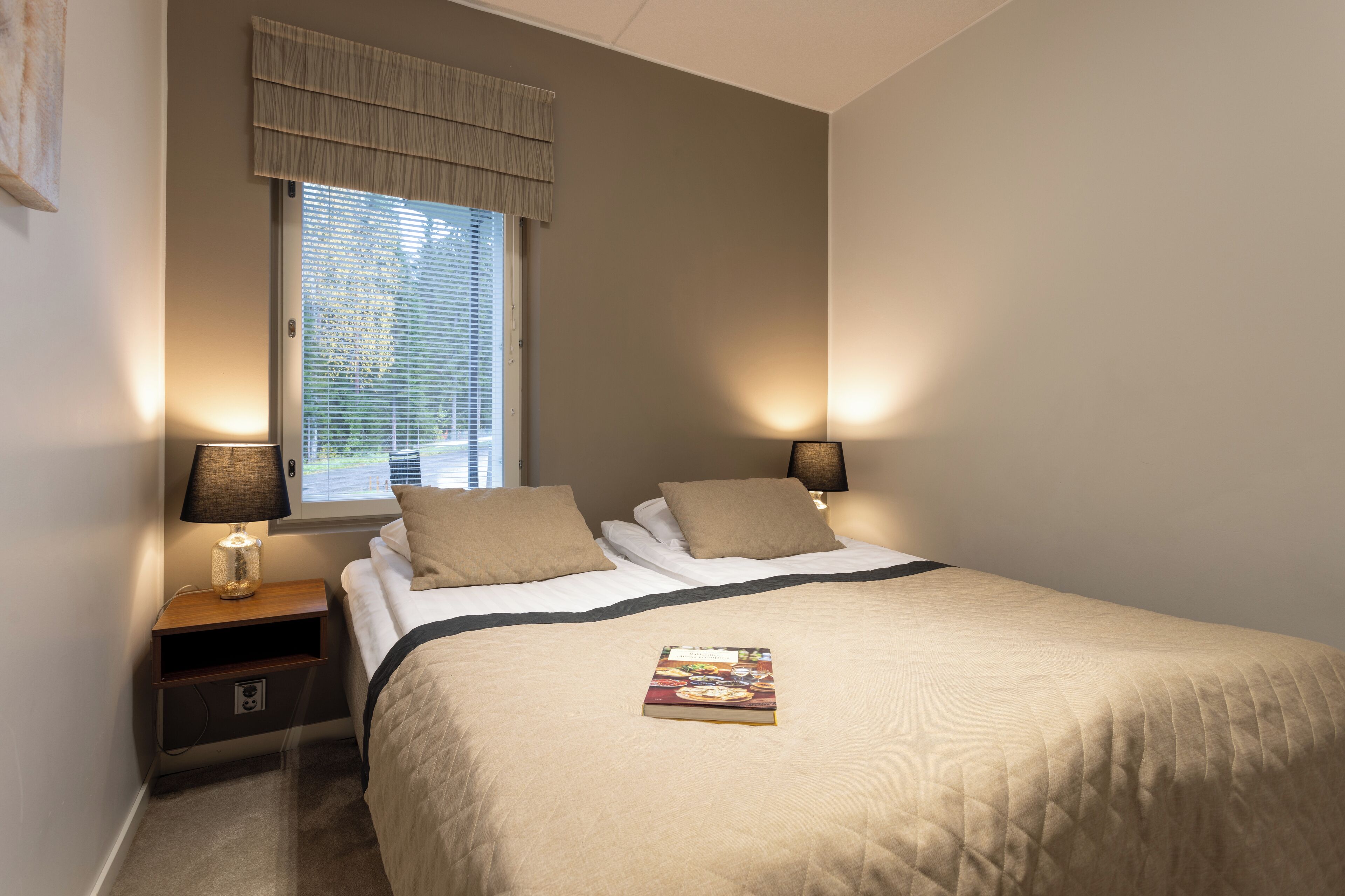 Een net opgemaakt bed met een boek erop, twee bedlampjes, een raam met jaloezieën en een nachtkastje met een lamp en een lade.