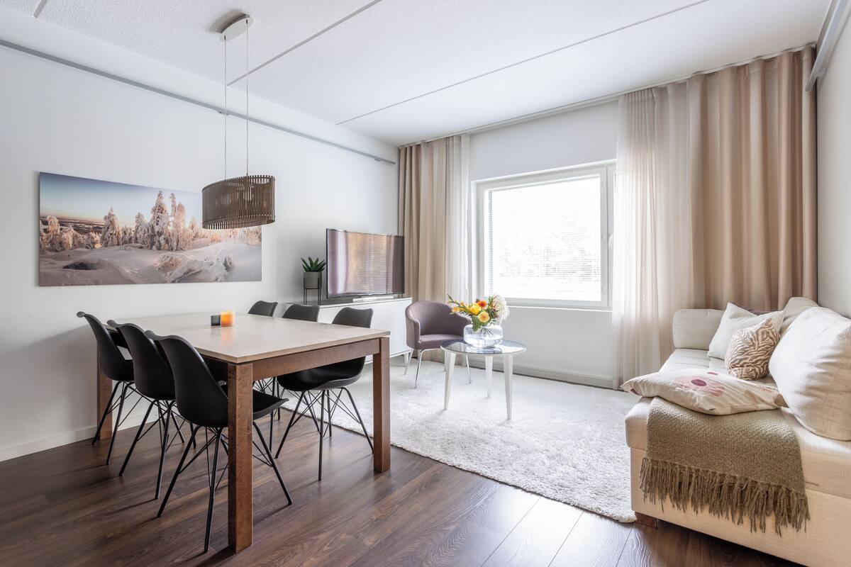 Ein modernes Wohnzimmer mit Essbereich, einer Couch, einem Fernseher und einem großen Fenster mit Vorhängen.