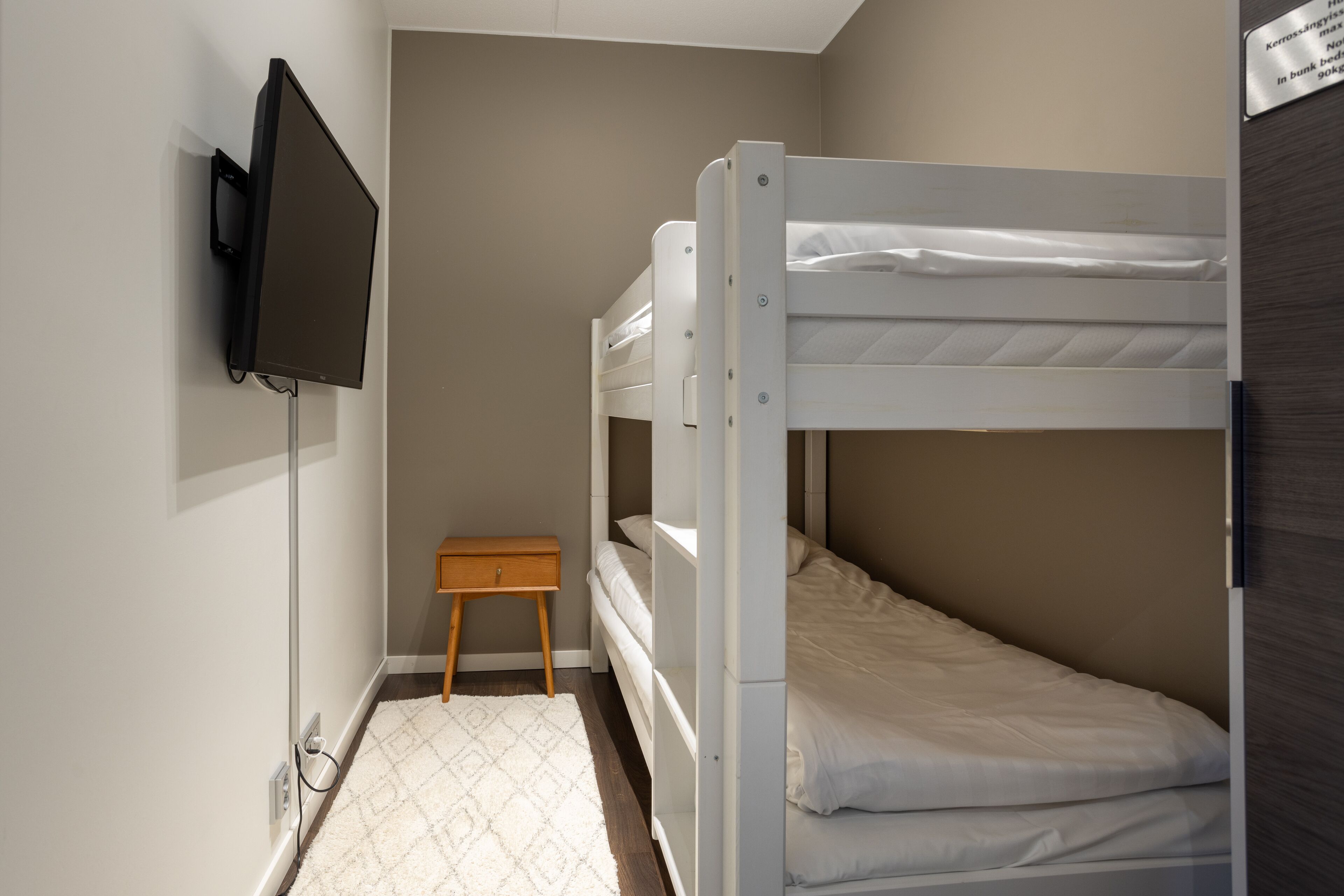 A bunk bed room with a television mounted on the wall, a small wooden nightstand, and a patterned rug on the floor.