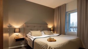 A neatly made bed with a quilted cover, two bedside lamps, a bedside table with a lamp, and a window with curtains. - Vuokatti Booking Suites   (Sotkamo)