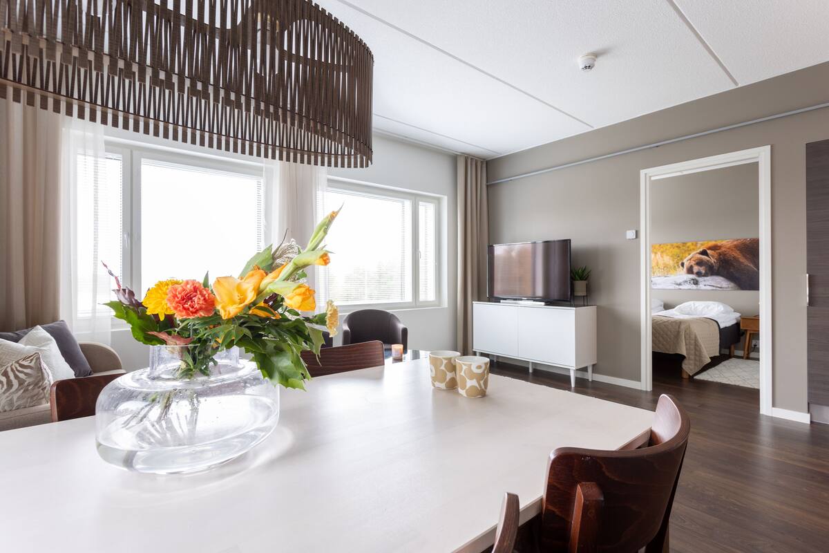 Ein modernes Wohnzimmer mit Esstisch, Stühlen, einem Blumenstrauß, einem Fernseher und einem Bett im Hintergrund.