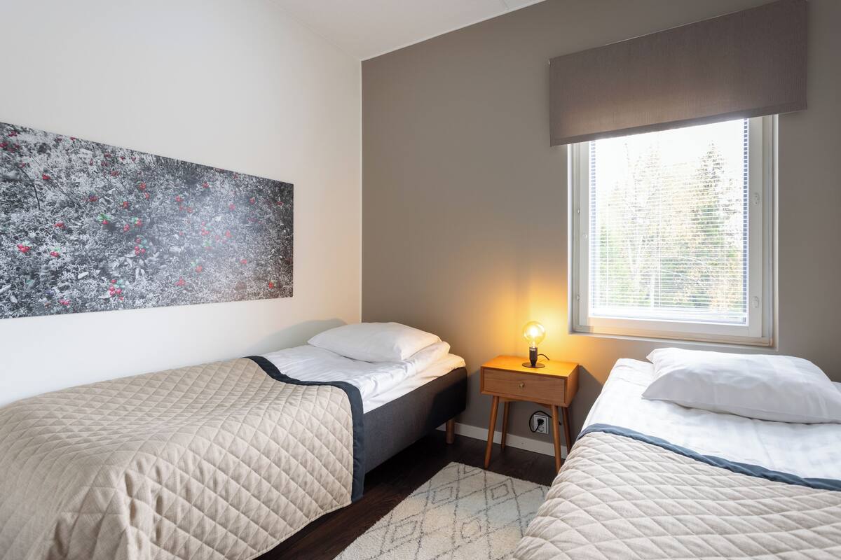 Ein Schlafzimmer mit zwei Betten, einem Nachttisch, einer Lampe, einem Fenster mit Jalousien und einem großen abstrakten Gemälde an der Wand.