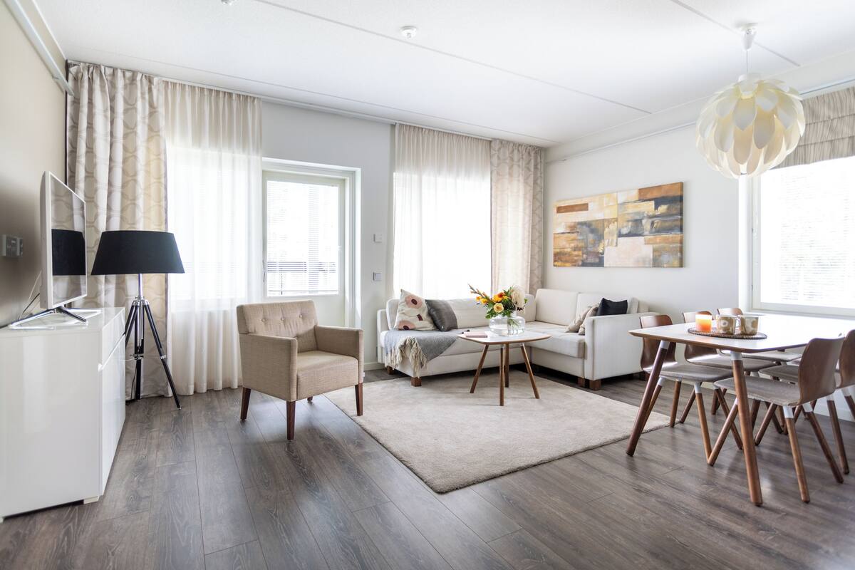 Ein modernes Wohnzimmer mit einer Couch, Sesseln, einem Couchtisch und einem Esstisch mit Stühlen. Es gibt eine Lampe, ein Bild an der Wand und ein Fenster mit Vorhängen.