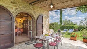 Un patio de piedra con una mesa y sillas, un área cubierta con techo, y vistas a un jardín con sillas y flores.