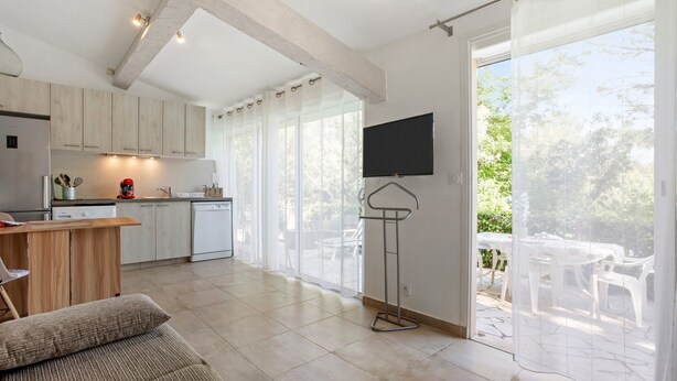 A modern kitchen with wooden cabinets, a dining area with a table and chairs, and a balcony with a view of greenery. - Studio Apartment 'Les Cyclamens - Les Oliviers' with Shared Pool, Wi-Fi and Air Conditioning (Saint-Raphaël)