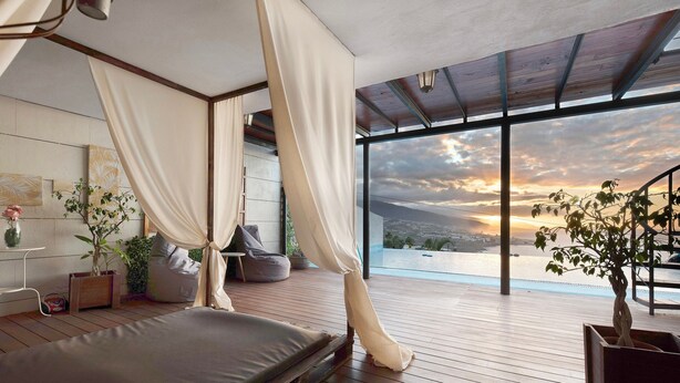 A bedroom with a bed, a large window, and a view of a sunset.