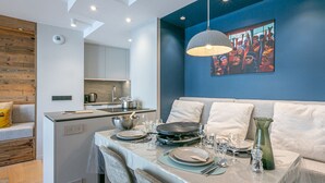 Un comedor moderno con una mesa dispuesta para cuatro, una cocina al fondo y una pared azul con un cuadro de colores.