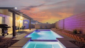 A rectangular swimming pool with a pergola and seating area on one side, surrounded by a brick wall and a landscaped garden. - Lavish 4BR Vegas Pool House w/ Heated Pool + Spa (North Las Vegas)
