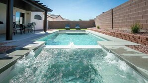 A swimming pool with a water feature, surrounded by a patio area with chairs and a table. - Lavish 4BR Vegas Pool House w/ Heated Pool + Spa (North Las Vegas)
