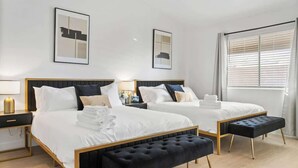A hotel room with two beds, a black bench, a bedside table with a lamp, and a window with blinds. - Lavish 4BR Vegas Pool House w/ Heated Pool + Spa (North Las Vegas)