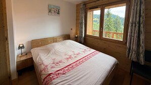 Ein Schlafzimmer mit einem Holzkopfende, einem Bett mit weiß-roter Bettdecke, einem Nachttisch mit Lampe und einem Fenster mit Blick auf Bäume und einen Balkon.