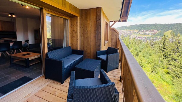 Eine Holzterrasse mit Gartenmöbeln, eine Glasschiebetür, die zu einem Essbereich im Innenbereich führt, und eine malerische Aussicht auf ein Dorf und Berge.
