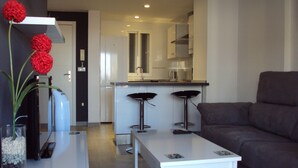 A modern living room with a white coffee table, a grey sofa, and two black bar stools. A kitchen with white cabinets and a stainless steel range hood is visible in the background. - Stunning 1 bedroom apartment with clear views across Benidorm (Benidorm)
