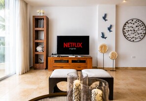 Ein Wohnzimmer mit Fernseher, der 'NETFLIX originals' zeigt, einem Holzregal, Wanddekoration und einem Fenster mit Vorhängen.
