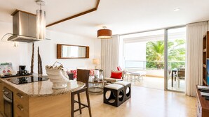 A modern kitchen with a central island, a dining area with a round table, and a balcony with a view of greenery.