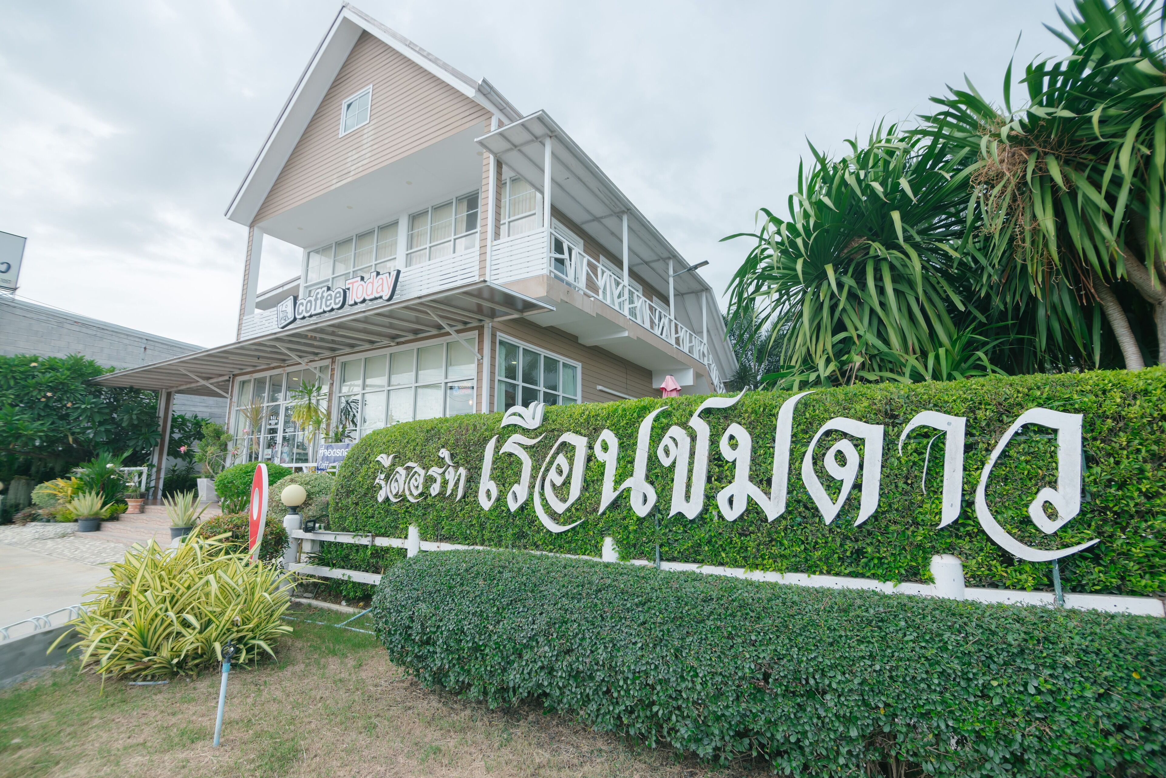 A two-story building with a balcony, surrounded by lush greenery and a sign with Thai script.
