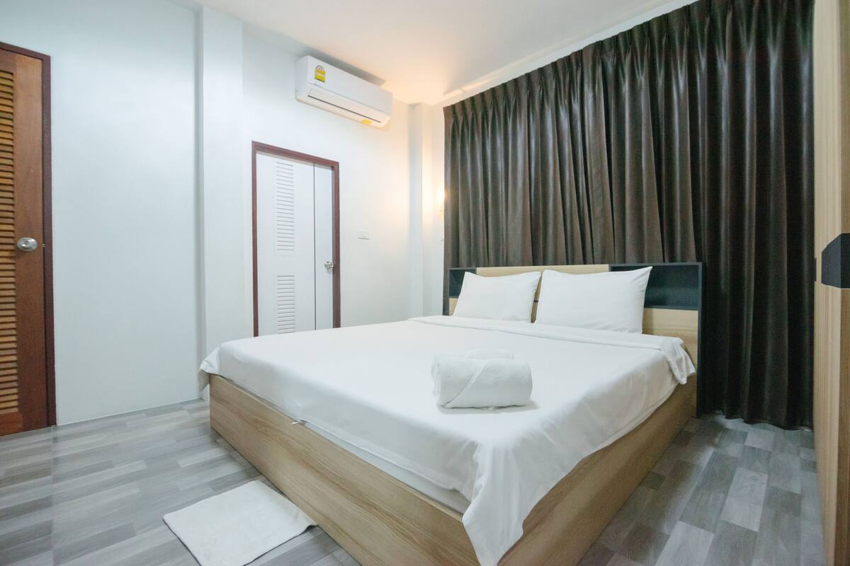A neatly made bed with white linens and a white towel, a wooden headboard, a white air conditioning unit on the wall, and a dark curtain covering a window.