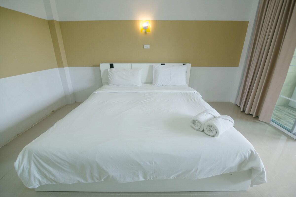 Una cama grande con ropa blanca, cabecera con lámpara y ventana con cortinas.
